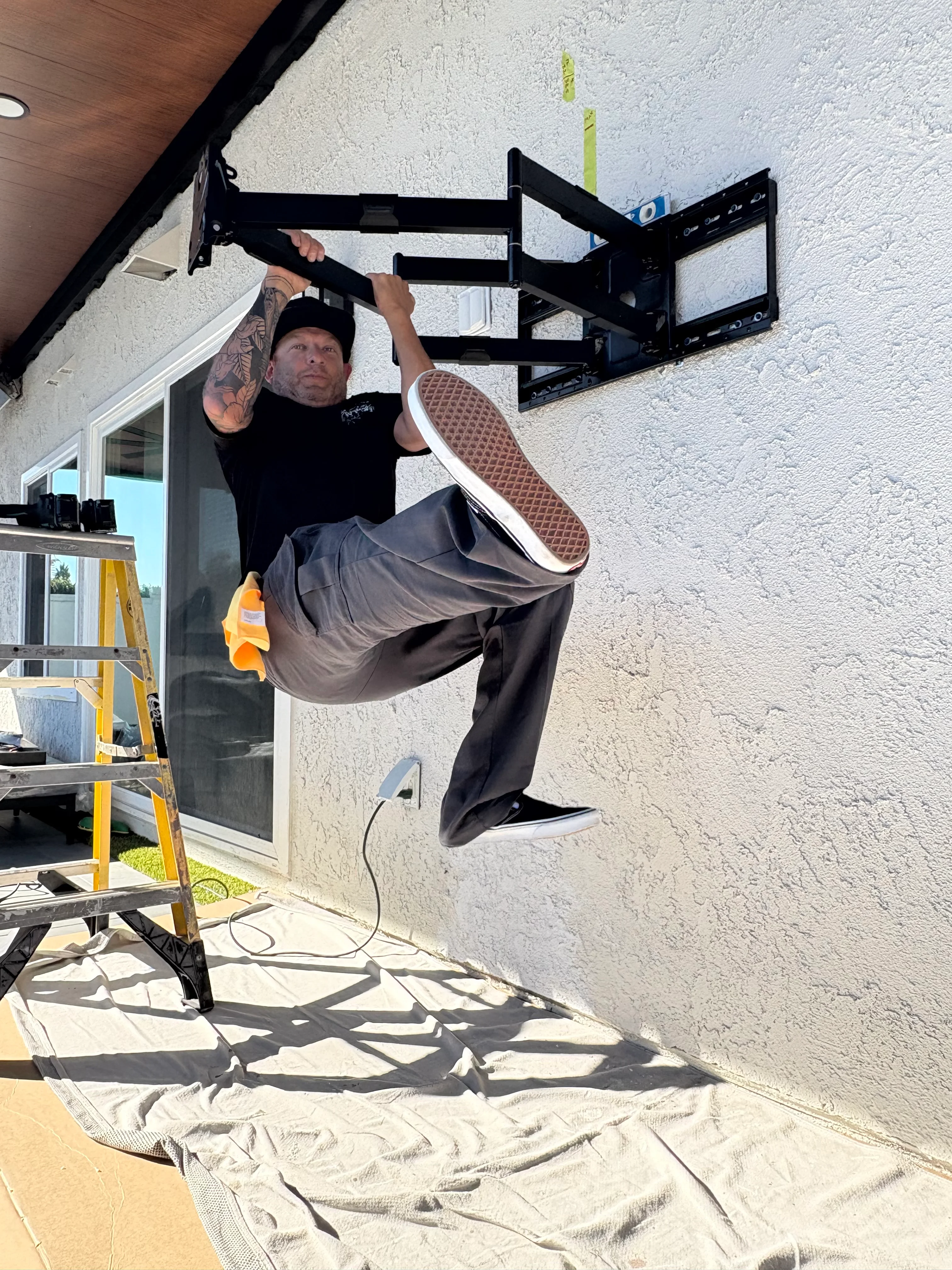 Technician installing black articulating TV mount on stucco wall with yellow ladder visible