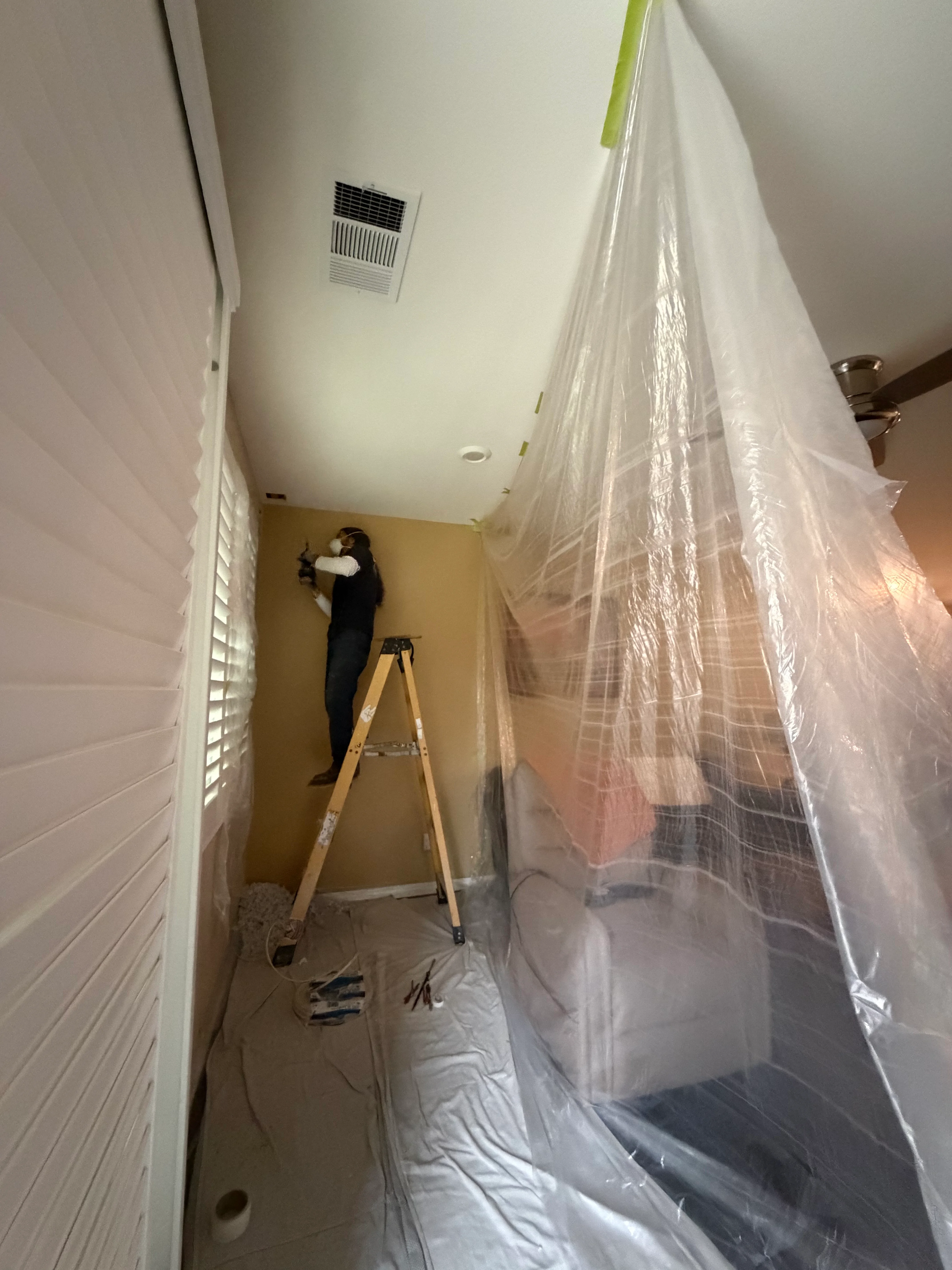Worker on ladder installing ceiling vent with plastic dust containment barriers in hallway